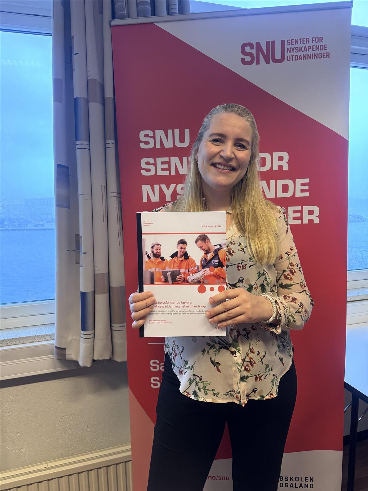 En smilende kvinne står foran et rødt SNU-banner og holder et diplom. Hun har på seg en lys, mønstret bluse og mørke bukser. - Klikk for stort bilde