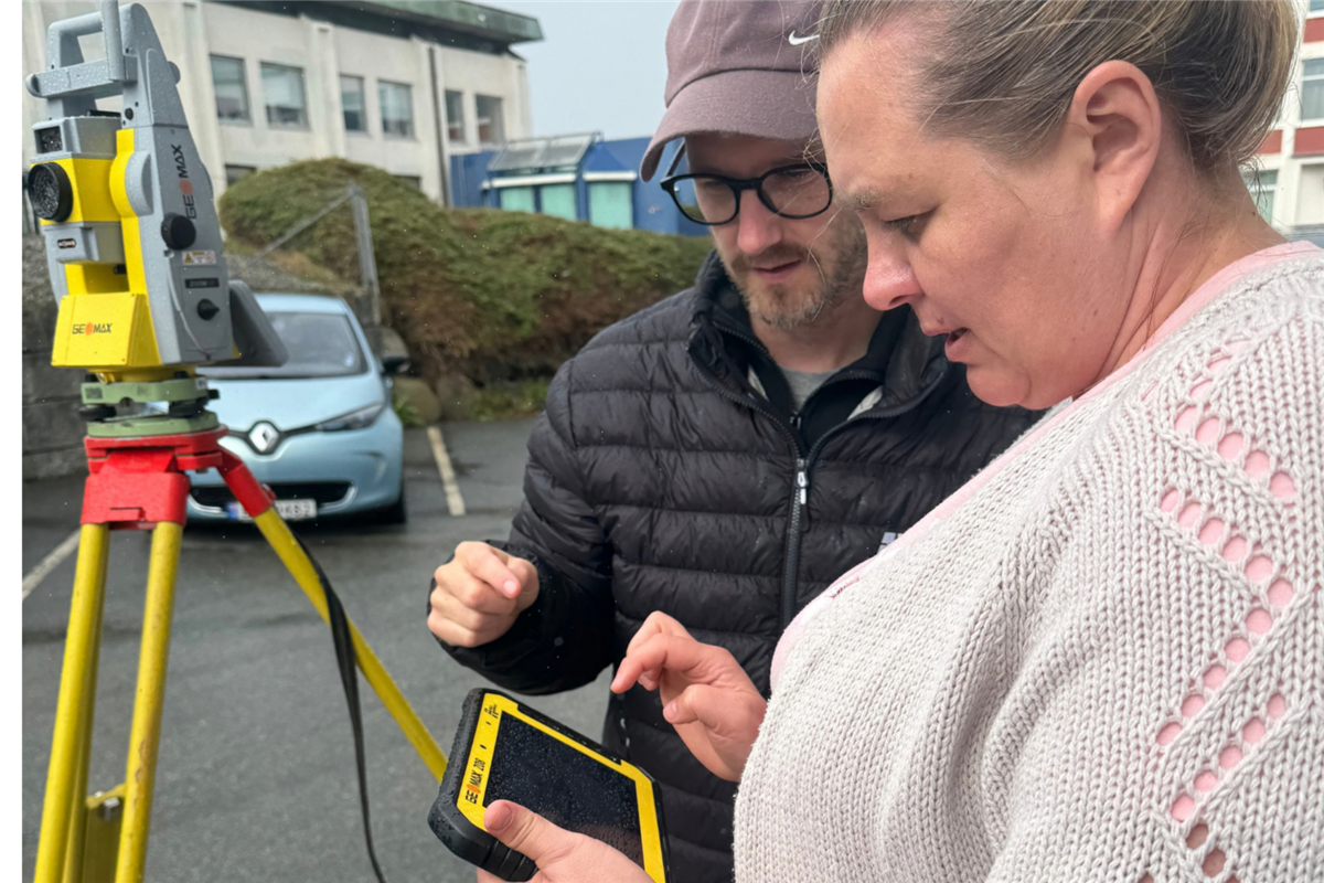 To personer bruker en gul og svart målekontroller ved siden av et landmålingsinstrument på et gult stativ. De står på en våt parkeringsplass utenfor en skolebygning. - Klikk for stort bilde